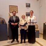 Trzy osoby stoją w muzeum, otoczone rzeźbami i obrazami. W centrum znajduje się popiersie osoby w mundurze. Ściany są jasne, a podłoga kremowa.