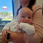 Na zdjęciu kobieta w różowej marynarce trzyma uśmiechnięte niemowlę w białym ubranku. W tle przez okno widać polską flagę i zielone drzewa. Obok na stole stoi bukiet kwiatów.
