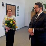 Na zdjęciu kobieta z bukietem żółtych i różowych kwiatów, obok mężczyzna w garniturze. Tło to jasne biuro z roślinami. Na szafce stoi zdjęcie.