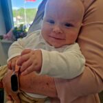 Na zdjęciu jest uśmiechnięte niemowlę na rękach dorosłego. W tle widać flagę Polski na maszcie i niebieskie niebo. Dziecko ma na sobie białe ubranko.