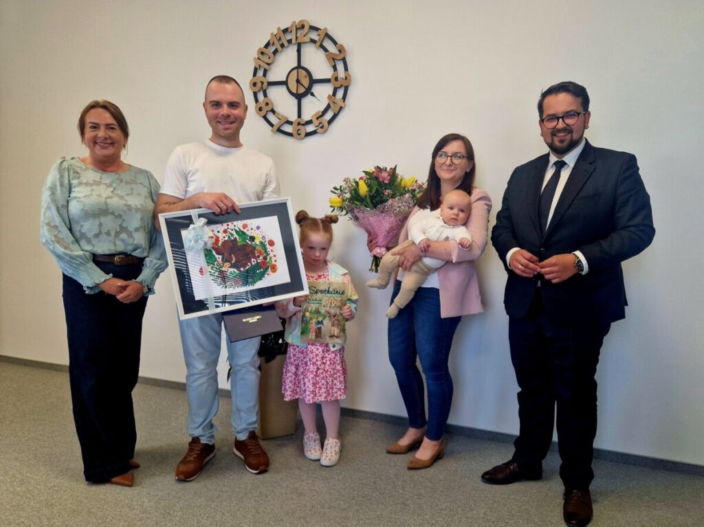 Grupa pięciu dorosłych i dwoje dzieci pozuje w formalnym otoczeniu. Osoba trzyma duży kolorowy obraz i tabliczkę, inna kwiaty. W tle jasna ściana z zegarem w stylu retro.