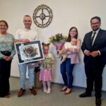 Grupa pięciu dorosłych i dwoje dzieci pozuje w formalnym otoczeniu. Osoba trzyma duży kolorowy obraz i tabliczkę, inna kwiaty. W tle jasna ściana z zegarem w stylu retro.