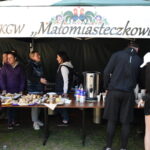 Stoisko KGW Małomiasteczkowi z ciastami. Pod zielonym namiotem, kilka osób w kurtkach. Na stole termos i kartony z napojami.