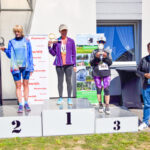 Trzy kobiety na podium z pucharami, w tle baner Mazowsze. Po prawej mężczyzna w kurtce. Ciepły, słoneczny dzień.