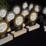 Trofea z profilowym wizerunkiem osoby, złote i srebrne detale. Stoją na czarnym tle. Nagrody na drewnianych podstawkach.