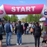 Na zdjęciu widać grupę ludzi stojących pod różowo-białym łukiem startowym. W tle zielone drzewa i niebieskie niebo. Atmosfera przypomina początek wydarzenia sportowego.