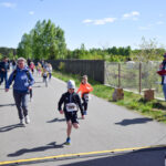 Na zdjęciu widać dzieci biegnące w wyścigu na wiejskiej drodze. W tle są drzewa i błękitne niebo. Widzowie stoją po bokach trasy.