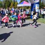 Grupa dzieci w kolorowych strojach rozpoczyna bieg na asfaltowej drodze. W tle tłum ludzi i biało-różowy namiot. Obok stoi duży, niebiesko-biały balon reklamowy.