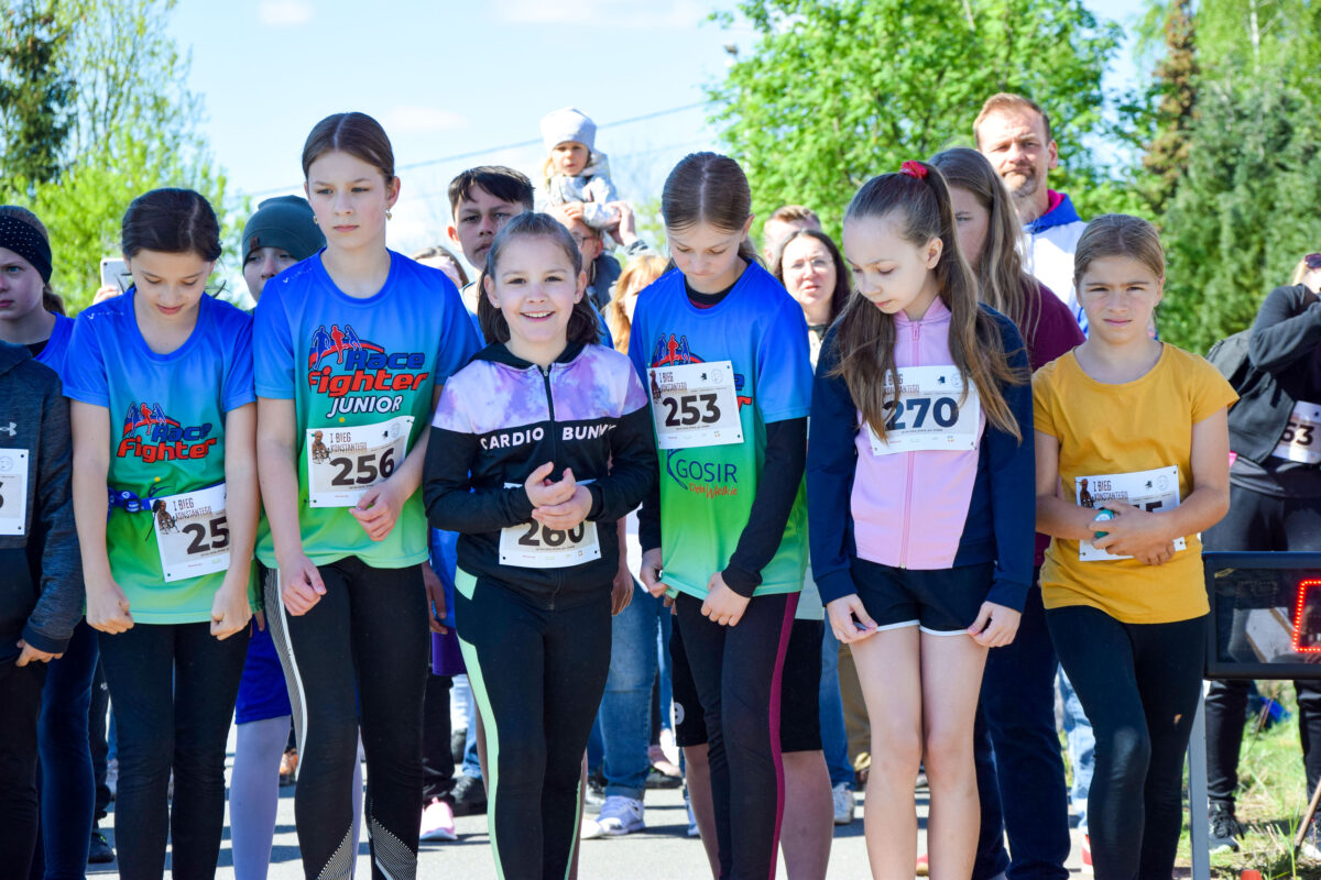 Grupa młodych biegaczy stoi na starcie w kolorowych koszulkach. W tle zielone drzewa i niebieskie niebo. Uśmiechy i determinacja na twarzach.