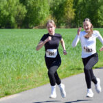 Na drodze biegną dwie dziewczyny w sportowych strojach. W tle zielone drzewa i trawa. Słoneczny dzień, numerki na koszulkach.