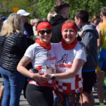 Na zdjęciu dwie uśmiechnięte kobiety w strojach do biegania, w białych koszulkach z kolorowymi wzorami. W tle tłum ludzi, część ubrana w kolorowe stroje sportowe. Sceneria jest jasna, a wokół znajdują się drz