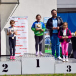 Dzieci stoją na podium z pucharami, w tle baner Mazowsze i stolik z trofeami, jasne tło budynku.