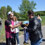 Na zdjęciu mężczyzna w różowo-czarnym stroju sportowym ściska dłoń drugiego mężczyzny w kurtce. W tle znajduje się zielone pole i grupa osób. Słońce świeci jasno, a niebo jest błękitne.