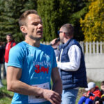 Biegacz w niebieskiej koszulce biegnie, w tle kibice w parku. Dzień słoneczny, zieleń i jasne kolory. Mężczyzna trzyma mikrofon.