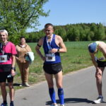 Trzech biegaczy stoi na drodze w słoneczny dzień. Ubrani w różne kolory koszulek i posiadają numery startowe. W tle zielone drzewa i błękitne niebo.