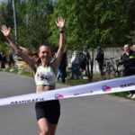 Kobieta przekracza metę maratonu z uniesionymi rękami, uśmiechnięta. Ma na sobie zieloną koszulkę i czarne spodenki. W tle widoczna jest zielona roślinność i kibice.