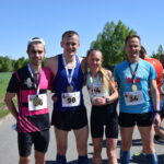 Czworo biegaczy pozuje z medalami na wiejskiej drodze. Są uśmiechnięci, ubrani w sportowe stroje. Tło stanowi zielone pole i drzewa.