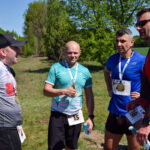 Na zdjęciu grupa mężczyzn w stroje sportowe z medalami, na tle zielonego lasu. Jeden daje znak kciuka w górę. Jasny dzień, błękitne niebo.