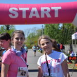Zdjęcie przedstawia dwie kobiety pod różowo-białą bramką startową biegu. Są uśmiechnięte i mają na sobie numery startowe. W tle widać innych uczestników oraz zielone drzewa.