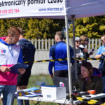 Pod namiotem odbywa się elektroniczny pomiar czasu. Mężczyzna w biało-niebiesko-różowej bluzie trzyma tablet. Tło stanowi tłum ludzi i drzewa.