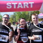 Trzy osoby w czarnych koszulkach z medalami stoją przed różowym banerem START. Za nimi zielone drzewa i niebieskie niebo. Tłum ludzi w tle.