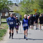 Na drodze odbywa się marsz Nordic Walking. Uczestnicy noszą numery startowe i stroje sportowe. W tle widać zielone drzewa i wiejską chatę.