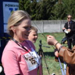 Nie wiem, kto jest na zdjęciu. Kobieta w różowej koszulce z numerem startowym, otrzymuje medal na zielonym tle, w tle ludzie i drzewa.