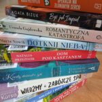 1000001222 Stos książek na stole z kolorowymi okładkami, w tle regały z książkami. Pomarańcz, czerń, biel i różne odcienie niebieskiego. Napisy na grzbietach wskazują tytuły i autorów.