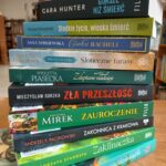 1000001223 Stos książek na drewnianym stole w bibliotece. Okładki w różnych kolorach, od zielonego po niebieski. W tle rozmyte regały z książkami.