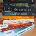 1000001224 Stos książek o różnobarwnych grzbietach leży na drewnianym stole. Każda książka ma inny kolor okładki i widoczny jest tytuł. Tło jest niewyraźne z półkami książek.