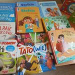 1000001225 Kolorowe książki dziecięce leżą na drewnianym stole. Okładki pełne są ilustracji i jaskrawych kolorów. Widać tytuły o rymowankach i pojazdach.