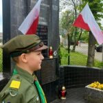 Chłopiec w harcerskim mundurze stoi przed pomnikiem z polskimi flagami. Monument jest ciemny, z białym napisem, tło zielone. Obok znajdują się znicze i kwiaty.