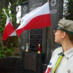 Pomnik upamiętniający, otoczony drzewami, z biało-czerwonymi flagami. Harcerz w mundurze stoi przed nim. Kwiaty i znicze u podstawy.