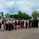 Ludzie stoją w szeregu na chodniku, jest pochmurne niebo. Harcerze w mundurach pośrodku. W tle drzewa i zabudowa miejska.