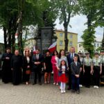 Grupa ludzi stoi przed pomnikiem w parku, w otoczeniu drzew. Mają formalne stroje, flagi Polski w tle. Tło to budynek i zielone drzewa.