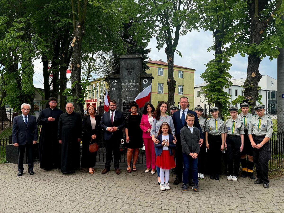 Grupa ludzi stoi przed pomnikiem w parku, w otoczeniu drzew. Mają formalne stroje, flagi Polski w tle. Tło to budynek i zielone drzewa.
