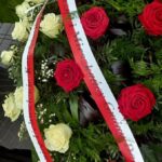 Wieniec z czerwonymi i białymi różami oraz zielonymi liśćmi. Na wieńcu biała wstęga z napisem. Tło to zieleń ogrodu.