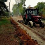 Traktor jedzie po nieutwardzonej drodze w lesie; ciągnie urządzenie do wyrównywania. W tle drzewa i zielone krzewy.