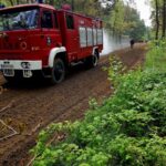 Czerwony wóz strażacki na drodze leśnej, z zielonymi drzewami po bokach. W tle strażak i lekki dym. Przód pojazdu widoczny z prawej strony.