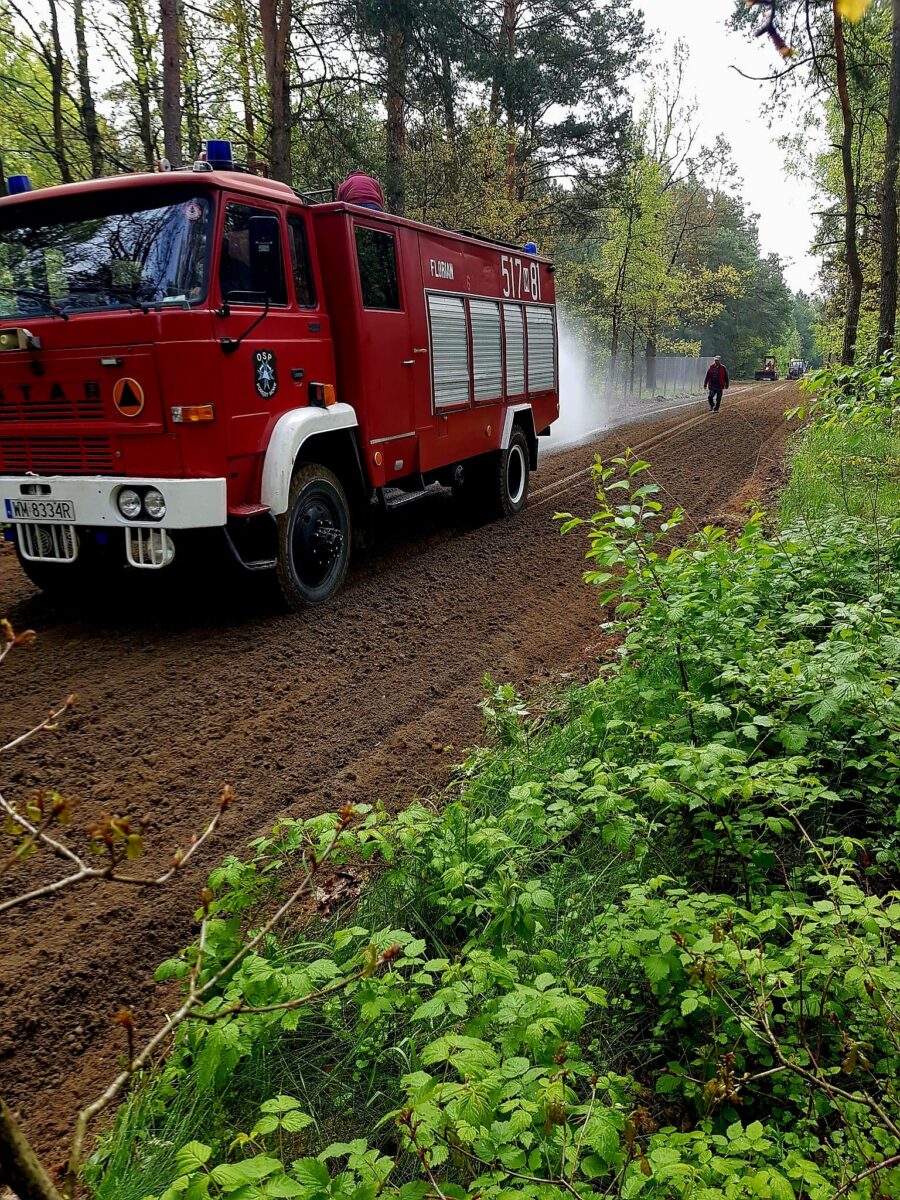 Czerwony wóz strażacki na drodze leśnej, z zielonymi drzewami po bokach. W tle strażak i lekki dym. Przód pojazdu widoczny z prawej strony.
