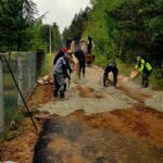 Grupa ludzi pracuje przy naprawie drogi, układając beton. W tle drzewa i chmurne niebo. Po lewej ogrodzenie.