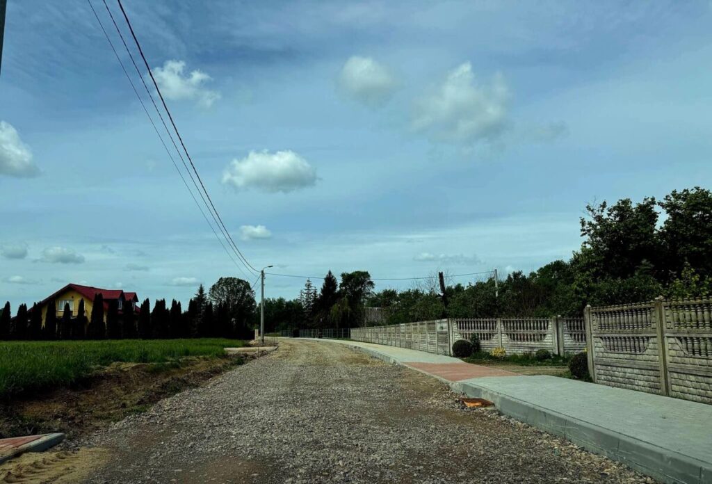 Droga żwirowa z chodnikiem prowadzi przez wiejską okolicę. Żółty dom z czerwonym dachem stoi pośród zieleni. Na niebie białe chmury.