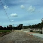 Droga żwirowa z chodnikiem prowadzi przez wiejską okolicę. Żółty dom z czerwonym dachem stoi pośród zieleni. Na niebie białe chmury.