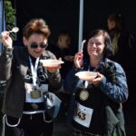 Dwie osoby jedzą zupę z plastikowych misek i łyżek. Mają medale na szyjach i numery startowe. W tle widać inne osoby.