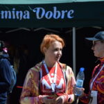 Trzy kobiety stoją przed namiotem z napisem Gmina Dobre. Jedna ma pomarańczową bluzę i medal, druga pije wodę. Kolory są jasne i żywe, tło ciemniejsze.