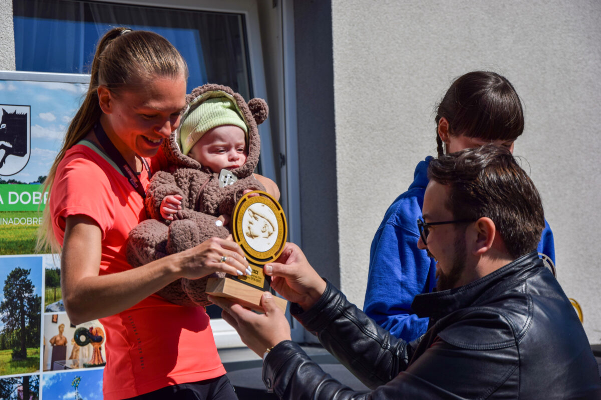 Kobieta w czerwonej koszulce trzyma dziecko w brązowym kombinezonie. Mężczyzna wręcza jej złoty medalion. Tło szare, na ścianie plakat.
