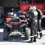 Strażacy w czarnych mundurach stoją obok czerwonego quada na brukowanym placu. W tle widać czerwony wóz strażacki. Obiekt znajduje się przed otwartym garażem.