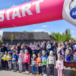 Na zdjęciu dzieci stoją pod różowo-niebieskim łukiem startowym. Mają kolorowe ubrania i numery startowe. W tle grupa dorosłych i zielone drzewa.