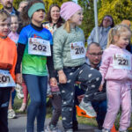 Na zdjęciu widać grupkę dzieci z numerami startowymi, przygotowujących się do biegu. Dzieci są ubrane w kolorowe stroje sportowe. W tle widoczni dorośli kibicują oraz wyświetlacz z czasem.