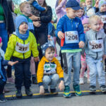 Grupa małych dzieci w kolorowych ubraniach stoi na linii startu. Mają numery startowe na torsach. W tle widoczni są dorośli i inne dzieci.